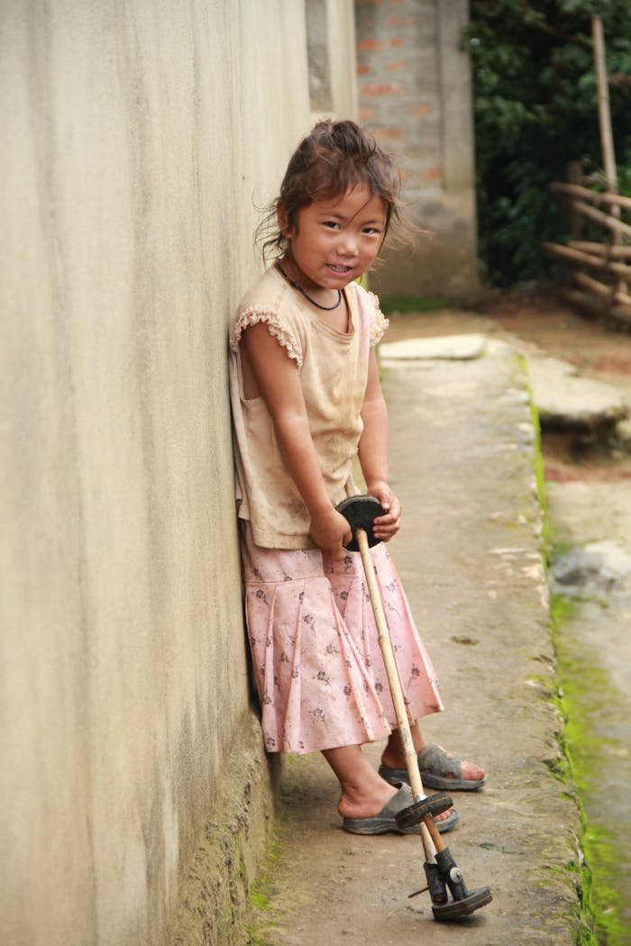 about-06 A young girl stands outdoors in a rural area, leaning on a staff with a shy smile.
