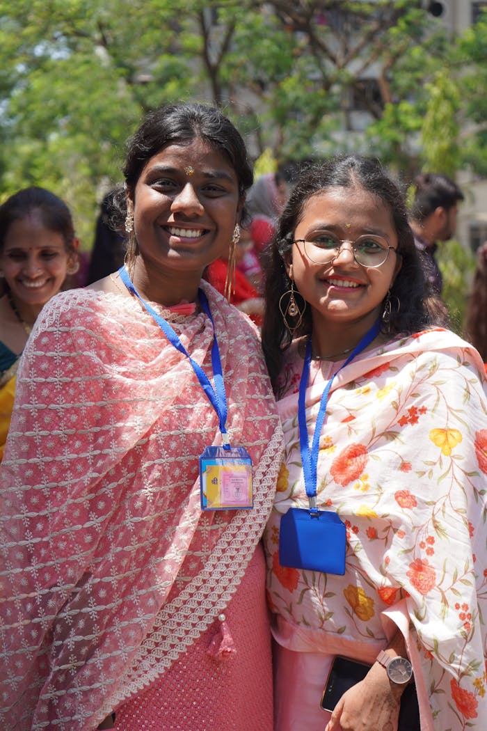Two young women in traditional clothing smiling outdoors, wearing floral sarees and ID badges.