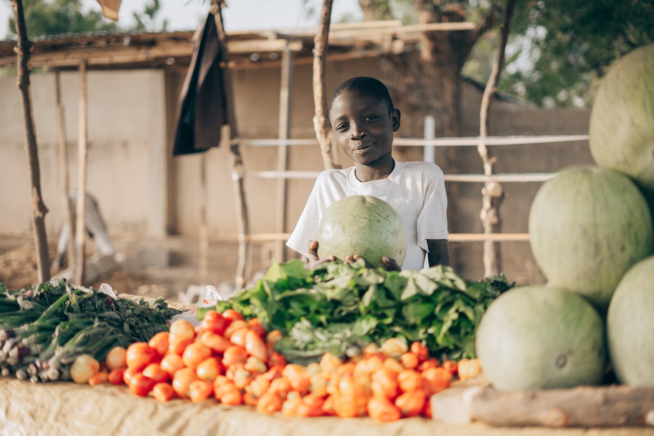 about-07 A young boy with fresh vegetables and watermelons at an outdoor market stall.